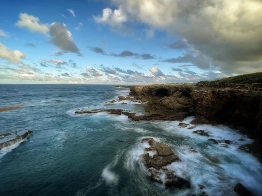 Coastal landscape featuring cliffs, ocean waves crashing on rocks, and a sky with scattered clouds during daylight in a scenic environment.