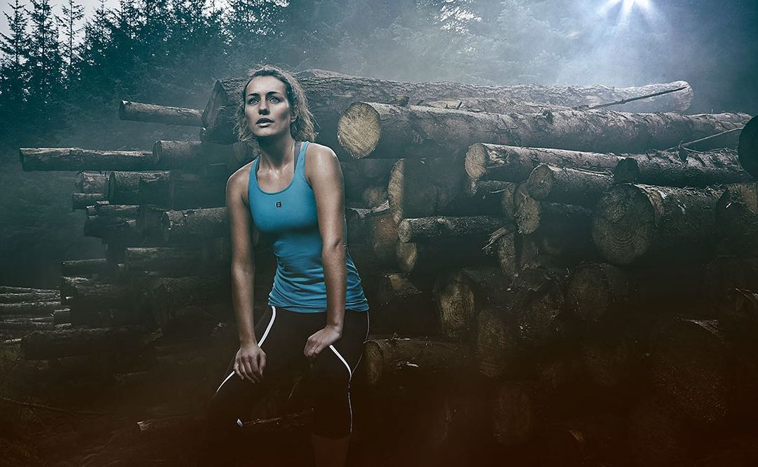 Female runner pausing in forest, resting hands on knees, wearing athletic wear with wood logs in the background and misty light.