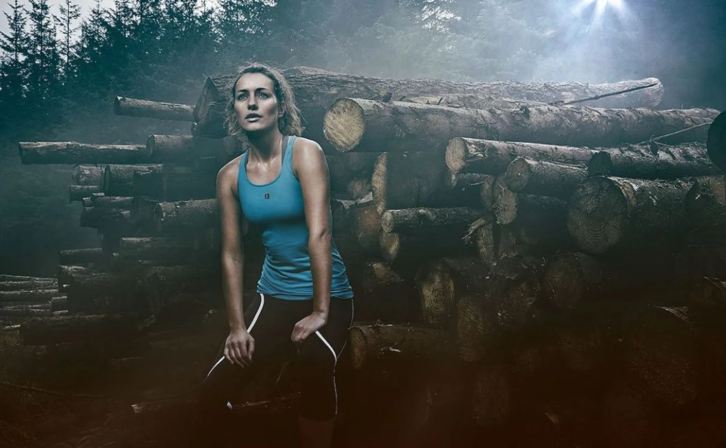Female runner pausing in forest, resting hands on knees, wearing athletic wear with wood logs in the background and misty light.