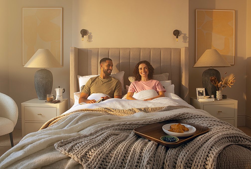 A man and woman are in bed with a tray of croissants and blueberries, beside end tables with lamps in a softly lit bedroom.