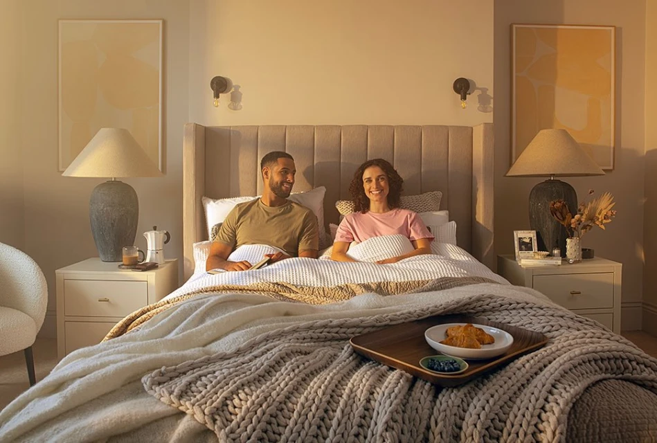 A man and woman are in bed with a tray of croissants and blueberries, beside end tables with lamps in a softly lit bedroom.