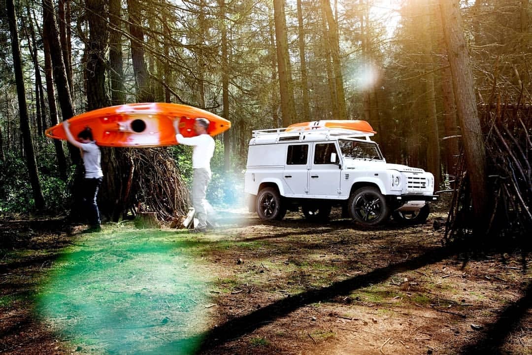 Two people carry an orange and white kayak near a white SUV in a forest with tall trees and dappled sunlight on the ground.