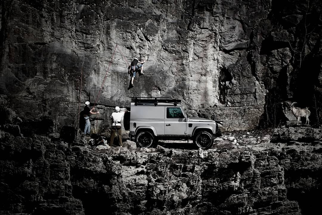 A silver SUV is parked near a rock climbing wall where a climber is ascending. Two adult males stand near the SUV, watching the climber.