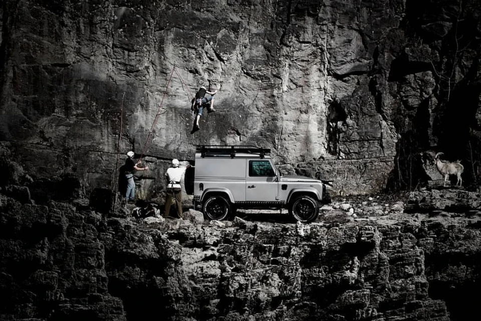 A silver SUV is parked near a rock climbing wall where a climber is ascending. Two adult males stand near the SUV, watching the climber.