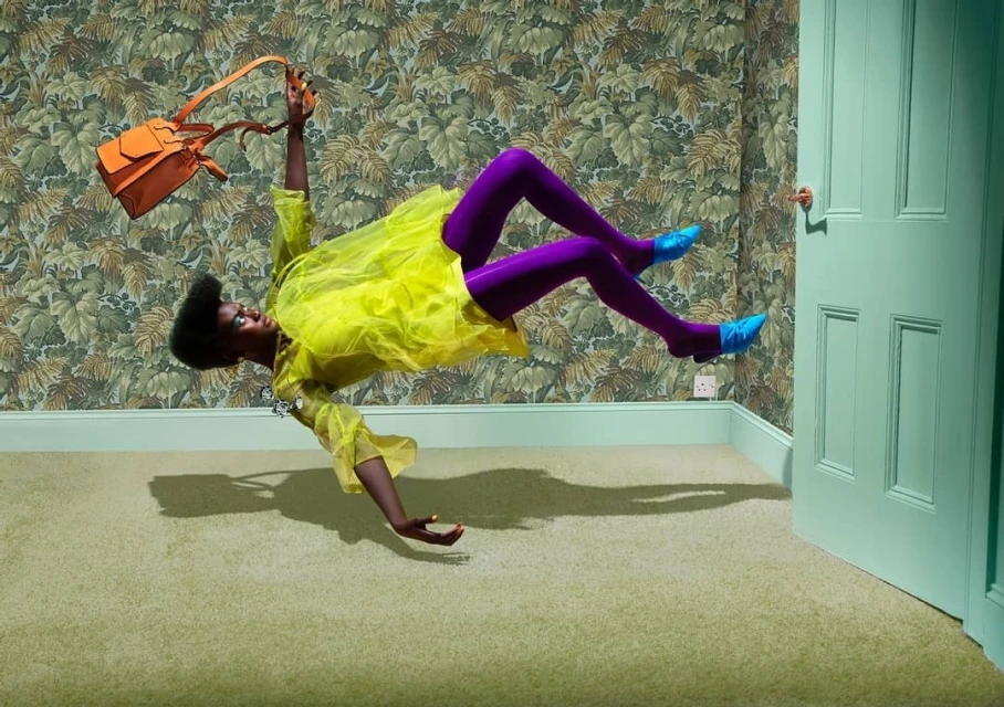 A Black female fashion model levitates indoors in a yellow dress, purple tights, and blue heels, holding an orange purse above her head.