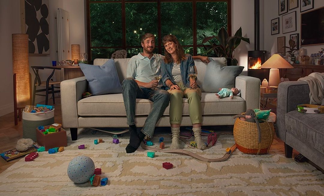 A couple, an adult man and an adult woman, sit on a couch in a living room with toys scattered on the floor, holding wine glasses.