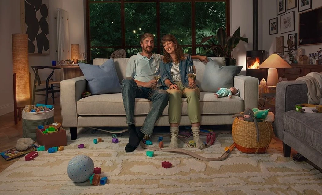 A couple, an adult man and an adult woman, sit on a couch in a living room with toys scattered on the floor, holding wine glasses.