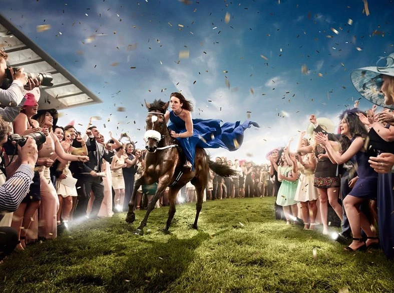 Female jockey riding a horse on a grass track, surrounded by crowds and photographers under confetti and a partly cloudy sky.