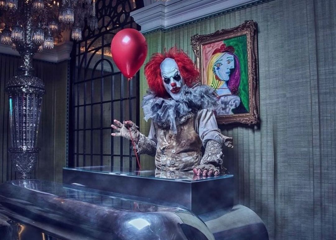 Clown in ornate interior poses with a red balloon next to modern art. Scary clown costume, ruff, red hair, and pale face paint.