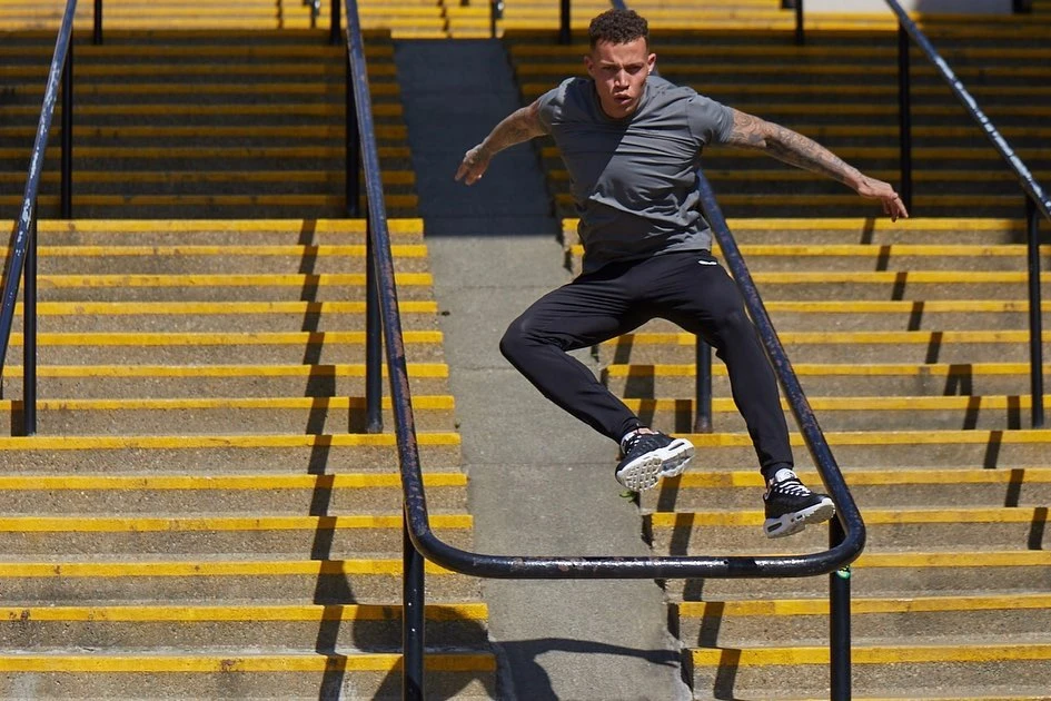 Athletic man in gray shirt and black pants jumps over black metal railing on yellow-striped stairs outdoors on a sunny day.
