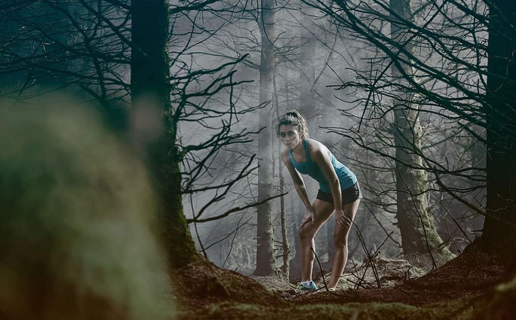 Athletic woman in running attire pauses to catch her breath during a trail run through a misty forest landscape.
