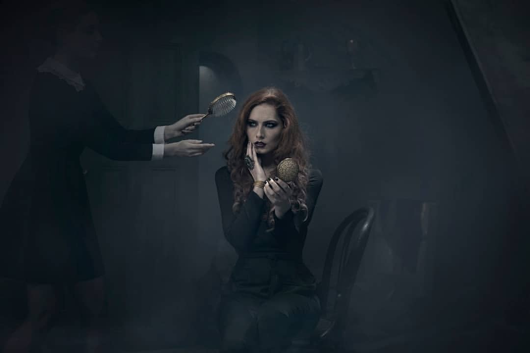 A woman with red hair sits in a chair and looks in a gold compact mirror while another person holds a hairbrush nearby.