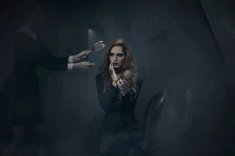A woman with red hair sits in a chair and looks in a gold compact mirror while another person holds a hairbrush nearby.