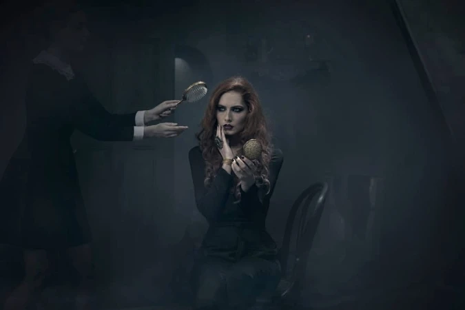 A woman with red hair sits in a chair and looks in a gold compact mirror while another person holds a hairbrush nearby.