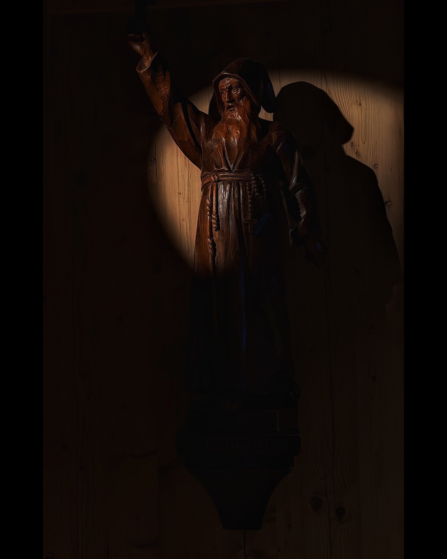 Dark, dramatic close-up of a wooden monk sculpture with a long beard, illuminated by a concentrated spotlight in a rustic indoor setting.