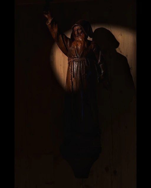Dark, dramatic close-up of a wooden monk sculpture with a long beard, illuminated by a concentrated spotlight in a rustic indoor setting.