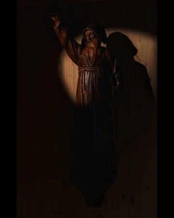 Dark, dramatic close-up of a wooden monk sculpture with a long beard, illuminated by a concentrated spotlight in a rustic indoor setting.