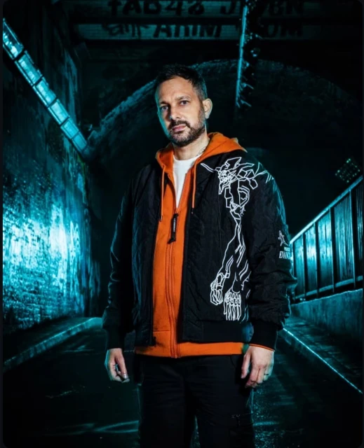 Man in black jacket and orange hoodie stands in dimly lit tunnel with brick walls and overhead pipes, creating a gritty urban scene.