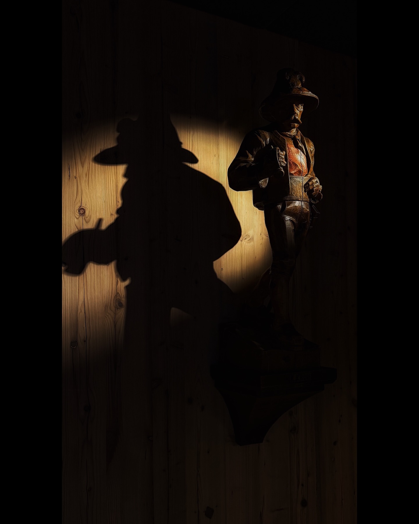 Wooden carving of a man in a hat and jacket is lit to cast a dramatic shadow on a wood-paneled wall, creating a vintage atmosphere.