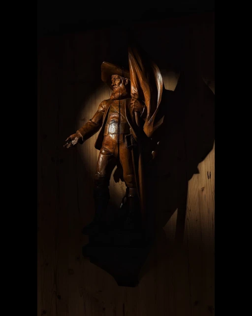 Detailed wood carving of a man in a hat and jacket with a cape, highlighted by a spotlight against a dark wooden background.