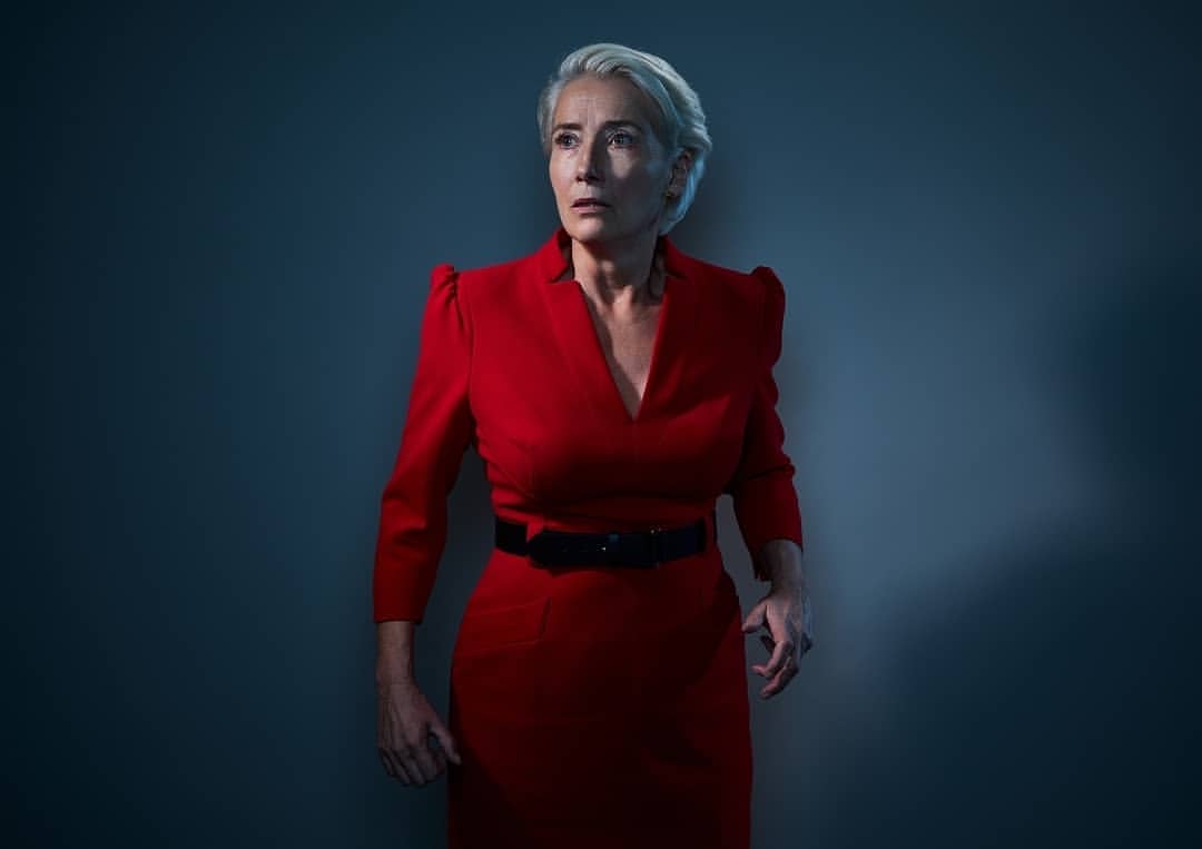 Emma Thompson poses in a red dress against a dim grey-blue background in a photo studio with soft directional lighting.