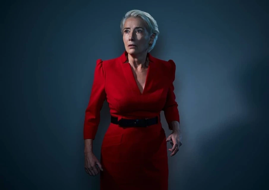 Emma Thompson poses in a red dress against a dim grey-blue background in a photo studio with soft directional lighting.