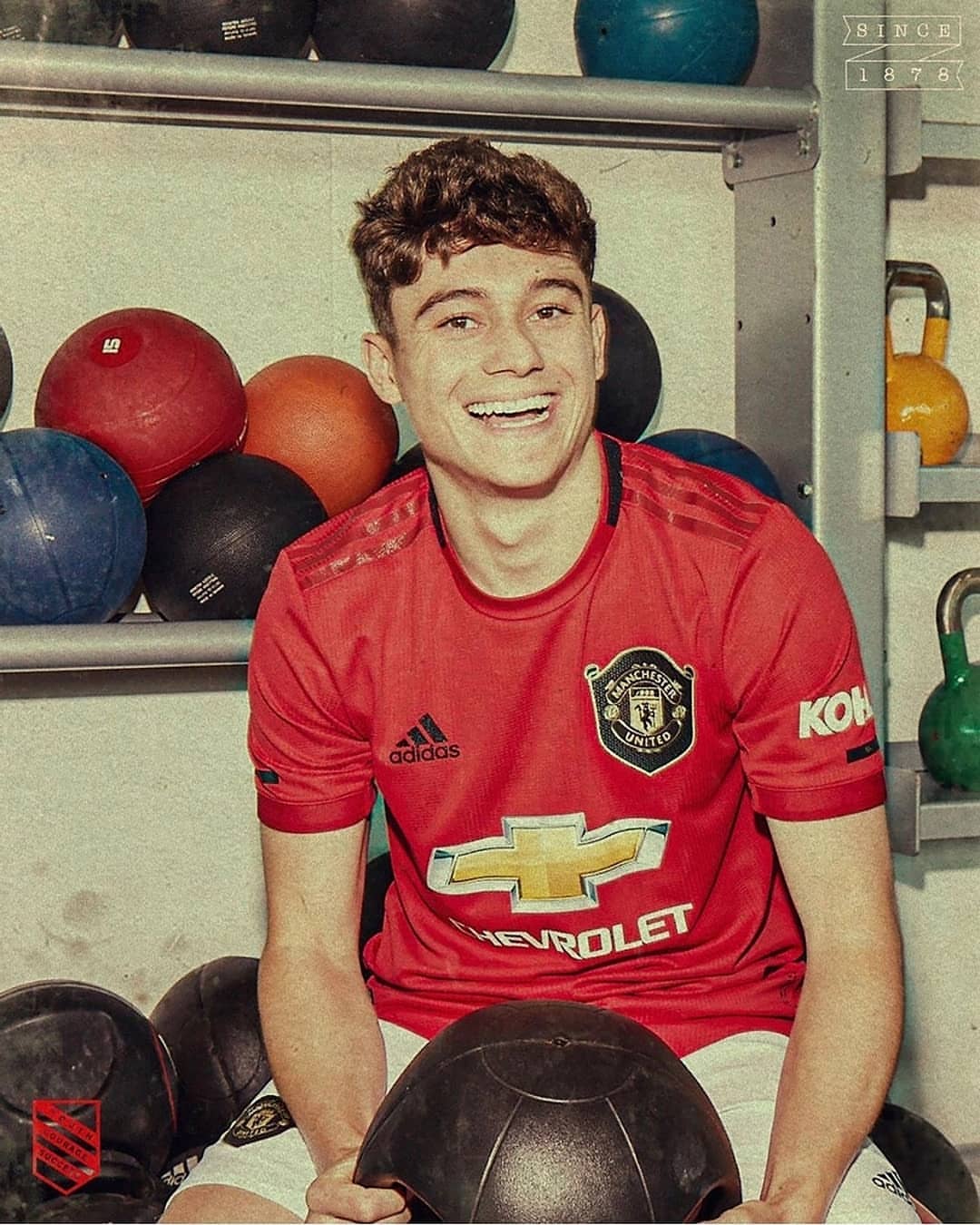Smiling soccer player in a red Manchester United jersey poses in a gym surrounded by black and red medicine balls on metal shelves.