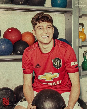 Smiling soccer player in a red Manchester United jersey poses in a gym surrounded by black and red medicine balls on metal shelves.