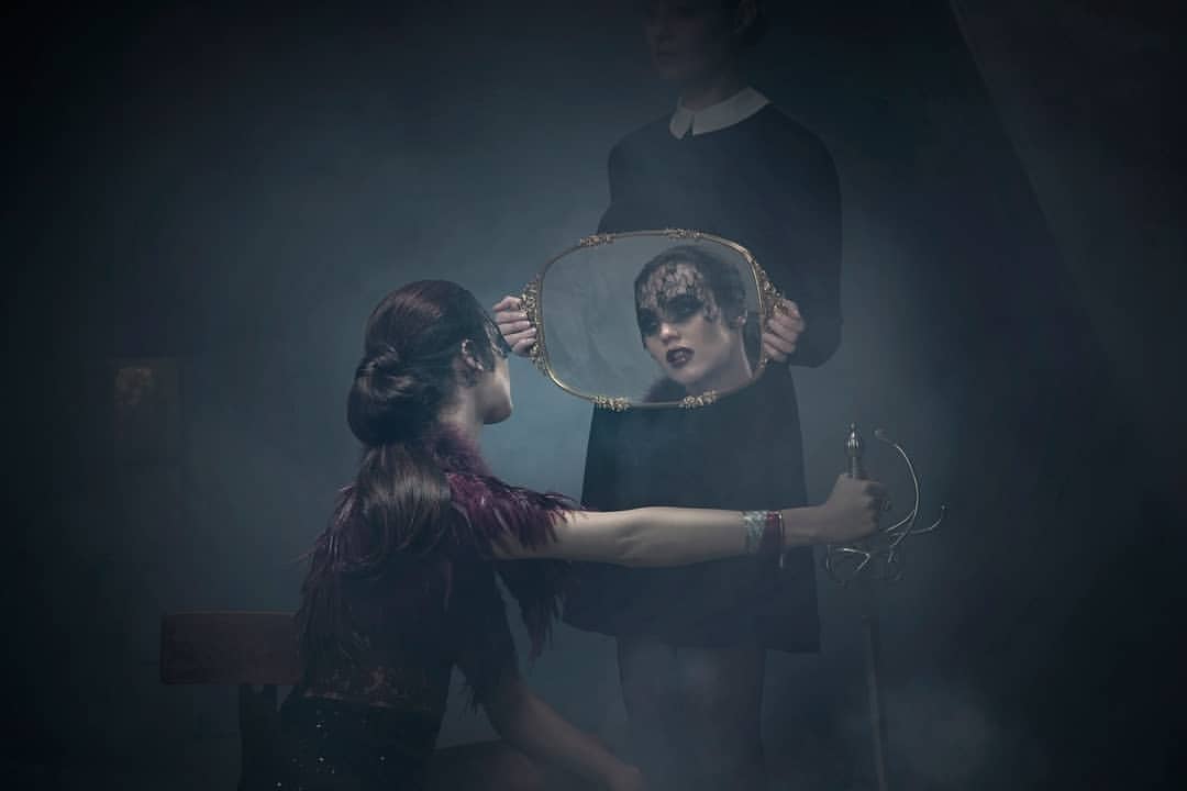 Artistic fashion photograph of a woman looking at her reflection in a mirror held by another woman in a dark, foggy indoor setting.