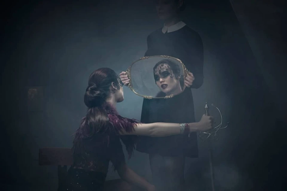 Artistic fashion photograph of a woman looking at her reflection in a mirror held by another woman in a dark, foggy indoor setting.
