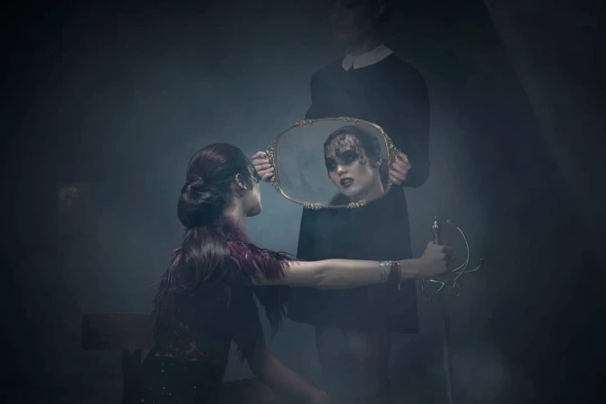 Artistic fashion photograph of a woman looking at her reflection in a mirror held by another woman in a dark, foggy indoor setting.