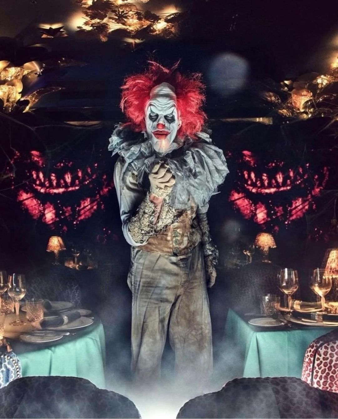 Creepy clown with red hair and white face paint stands behind a table set with plates and wine glasses in a dimly lit room.