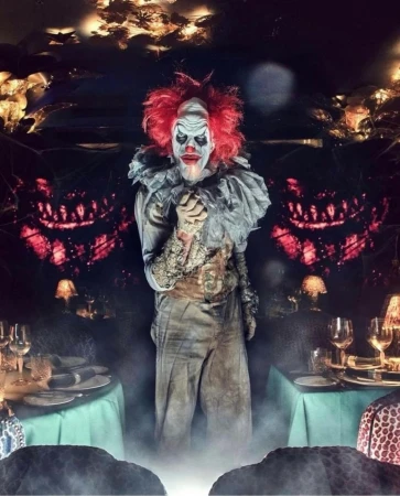 Creepy clown with red hair and white face paint stands behind a table set with plates and wine glasses in a dimly lit room.