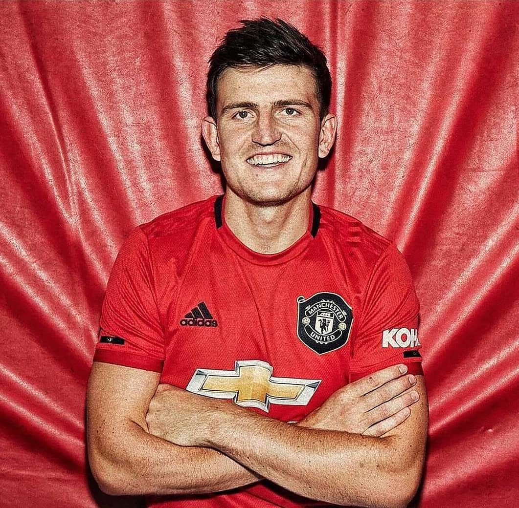 A waist-up studio shot of a smiling man in a red Manchester United jersey with arms crossed, set against a textured red backdrop.