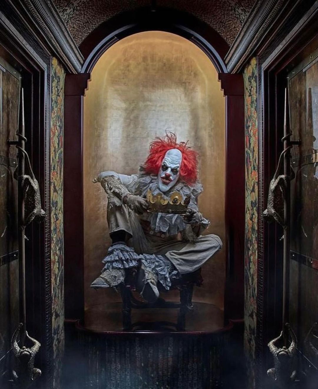 Creepy clown with red hair and white face paint sits holding a gold crown against a gold backdrop in a dark, interior scene.