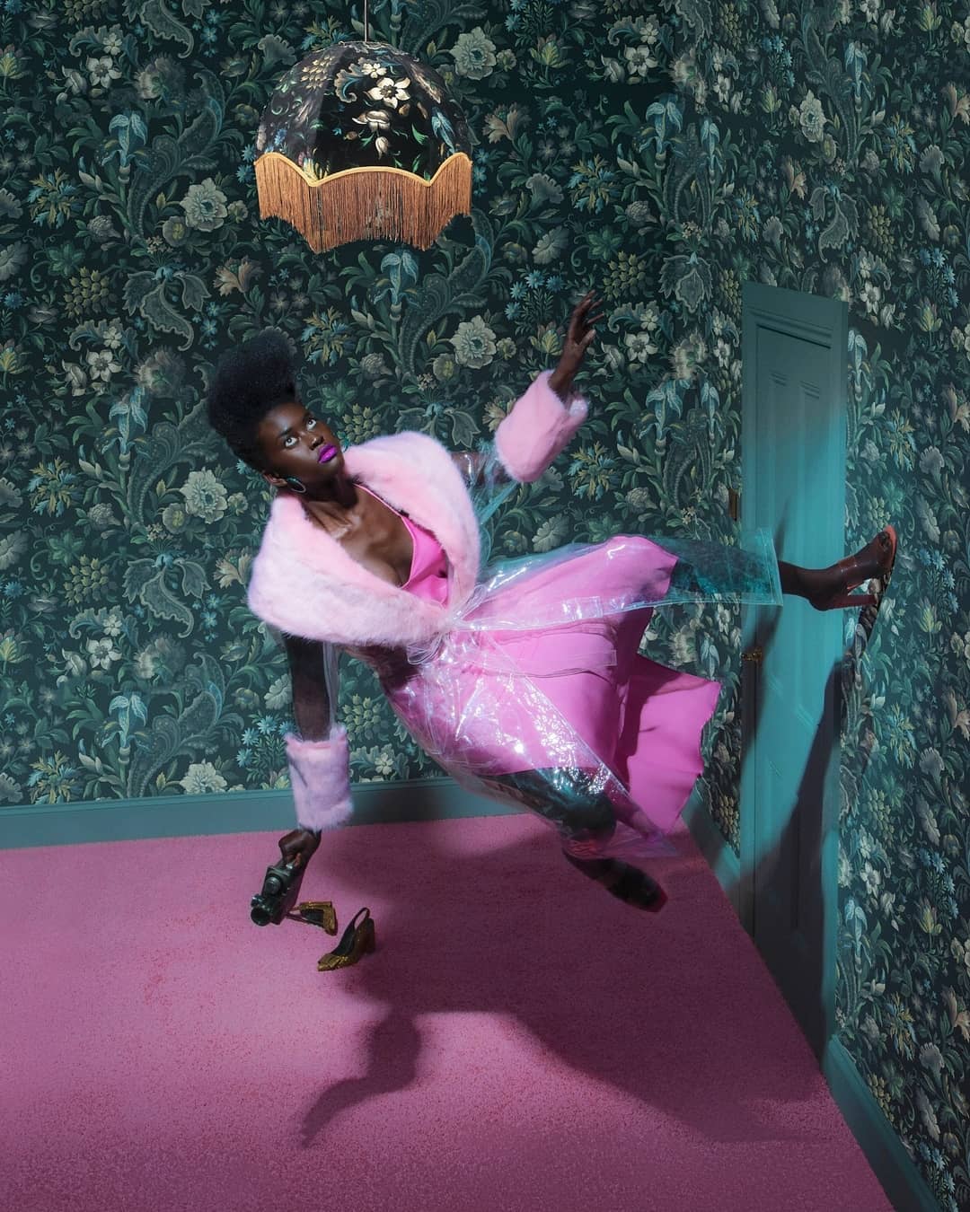 A dark-skinned woman in pink fashion levitates indoors, wearing a coat and dress, with shoes on the floor and floral wallpaper.