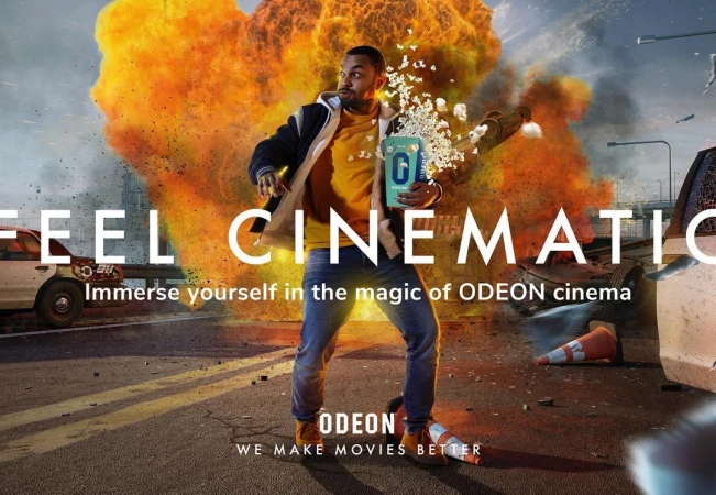 An adult male stands on a street holding popcorn with an explosion behind him in an advertisement for ODEON cinema movies.