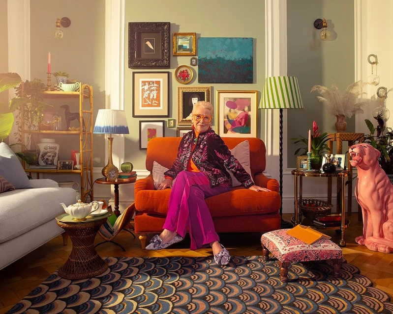 Stylish senior woman with short white hair sitting in an orange armchair in a light green-walled living room filled with art and decor.
