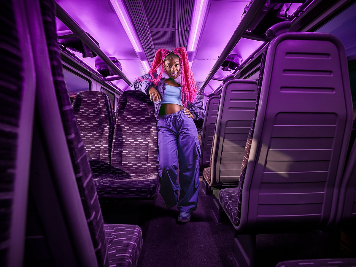 Stylish young woman with pink hair in a train car, wearing a blue crop top, purple pants, and a jacket, illuminated by purple lighting.