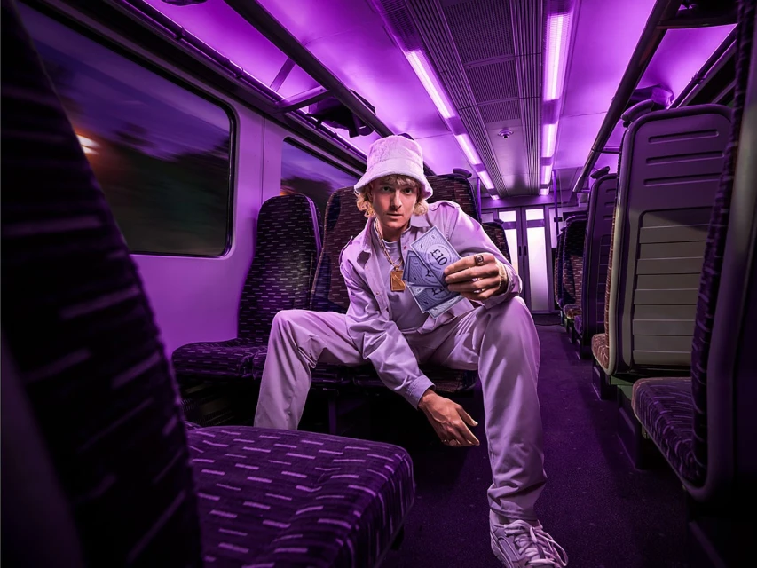 Young adult poses inside train car, holding money, wearing matching lilac bucket hat, pants, and jacket illuminated by purple lighting.