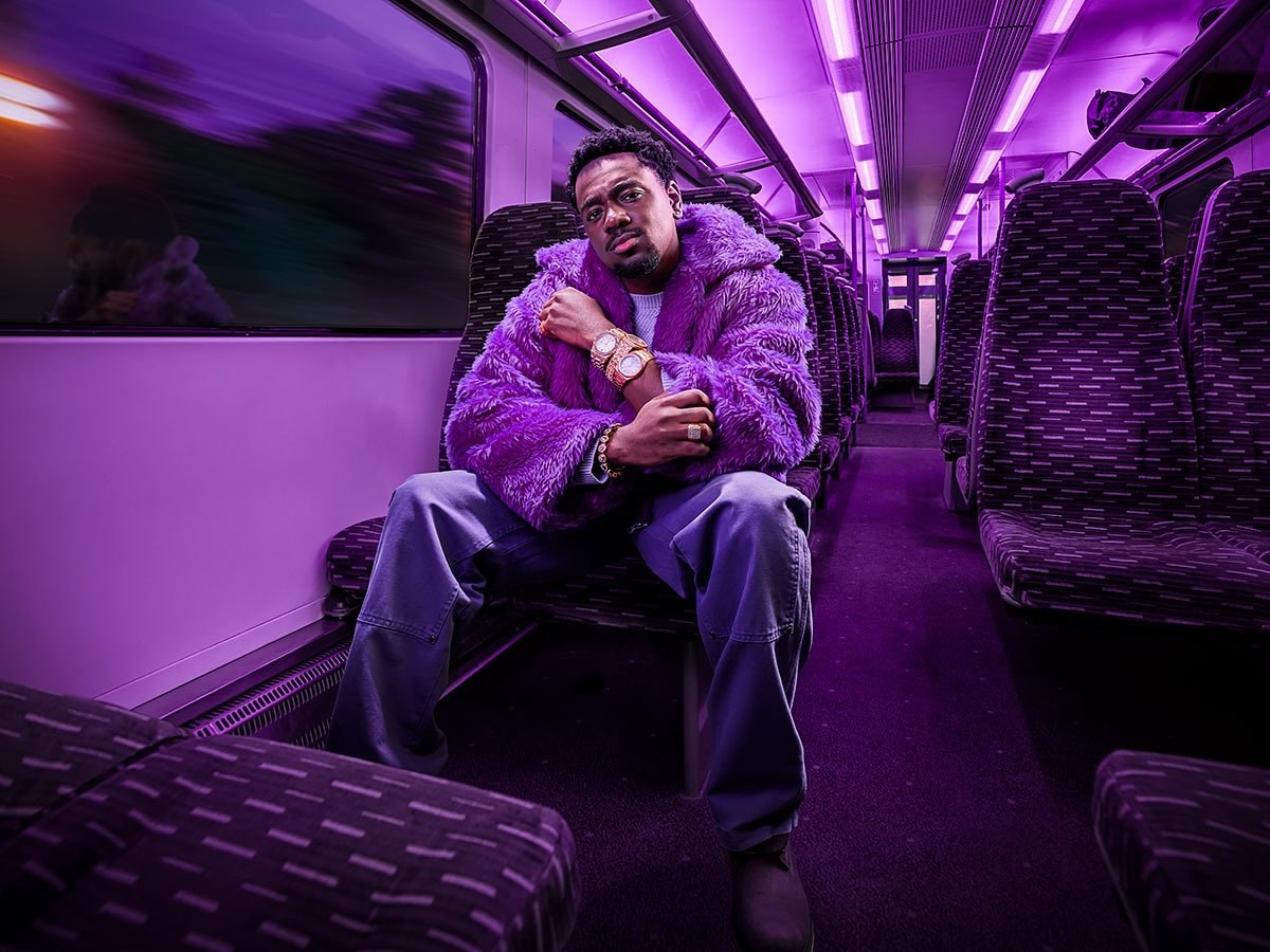 A man with a beard poses in a purple faux fur coat, jeans, and boots inside a train car with purple lighting and patterned seats.