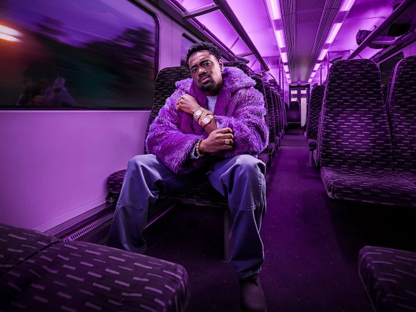 A man with a beard poses in a purple faux fur coat, jeans, and boots inside a train car with purple lighting and patterned seats.