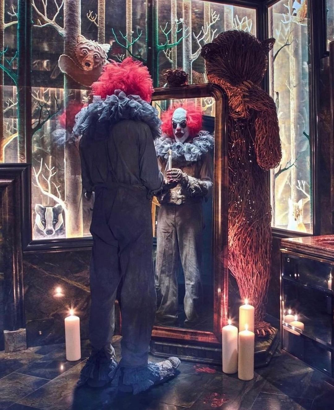 Clown with red hair and dark costume stands before a mirror reflecting his painted face, surrounded by candles in a dimly lit room.