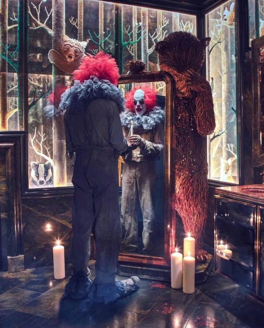Adult in clown costume looks in mirror, reflecting clown face, set indoors with candles and animal artwork, creating eerie atmosphere.