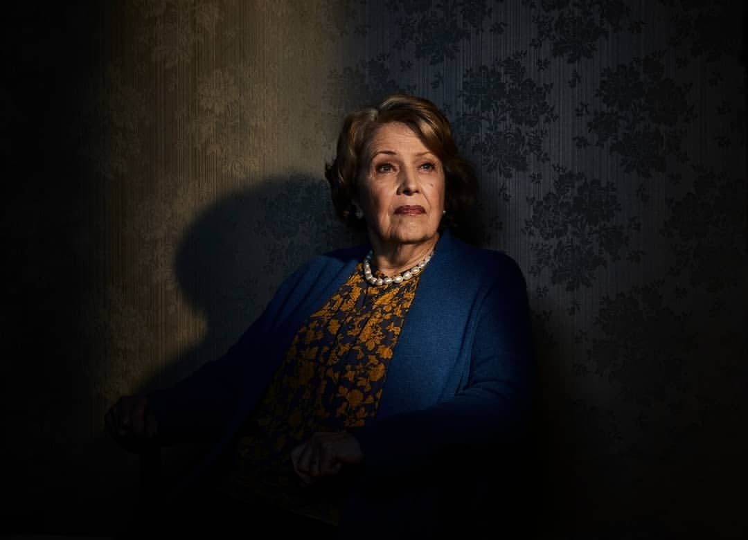 Elegant elderly woman in a patterned dress and blue cardigan poses indoors against a floral damask wallpaper, looking off-camera.