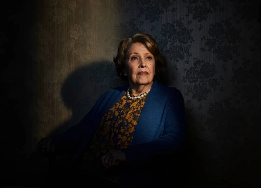Elegant elderly woman in a patterned dress and blue cardigan poses indoors against a floral damask wallpaper, looking off-camera.