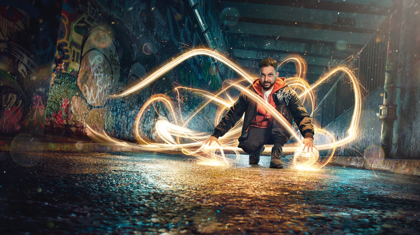 A man is crouched in a tunnel with graffiti, seemingly creating light patterns with his hands in a dynamic, visually striking composition.