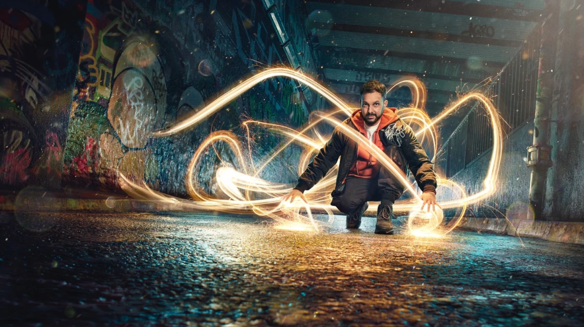 A man is crouched in a tunnel with graffiti, seemingly creating light patterns with his hands in a dynamic, visually striking composition.