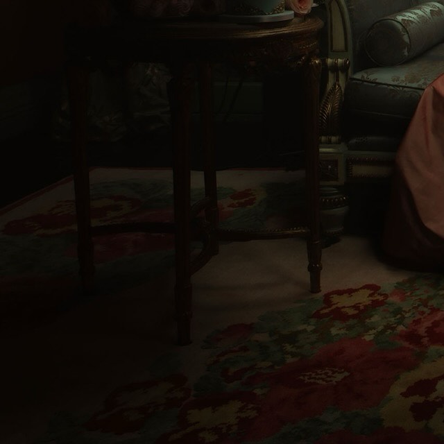 Dark interior scene featuring a vintage floral rug, an ornate table with floral decor, and a partially visible antique chair.