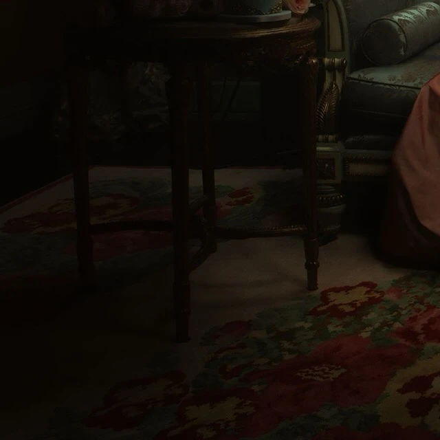 Dark interior scene featuring a vintage floral rug, an ornate table with floral decor, and a partially visible antique chair.
